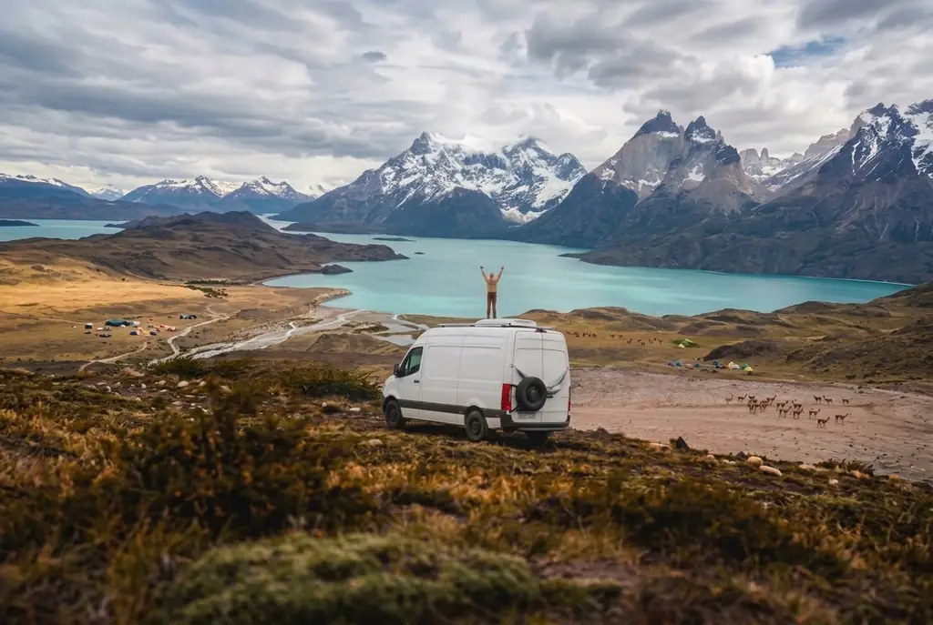 Van campista no alto de um morro na Patagônia, com uma pessoa de braços abertos sobre o teto, guanacos na estepe, um lago turquesa e montanhas nevadas ao fundo